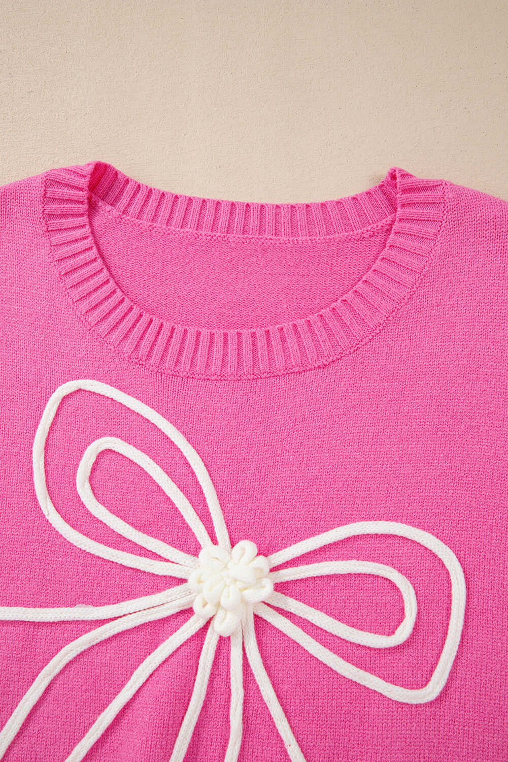 Soft pink sweater with a white corded bow design and ribbed collar, featuring a close-up of the knit texture.