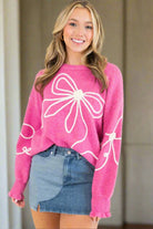 Woman wearing a soft pink sweater with corded bows and ruffled cuffs, styled with a denim skirt, showcasing trendy and comfortable fashion.