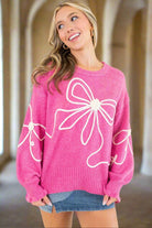 Pink sweater with corded bows design, dropped shoulder, and ruffled cuffs worn by smiling woman in a stylish indoor setting.