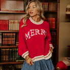 A model wearing the MERRY MOMENTS SWEATER, a cozy red knit sweater with a festive 'MERRY' graphic and relaxed fit.