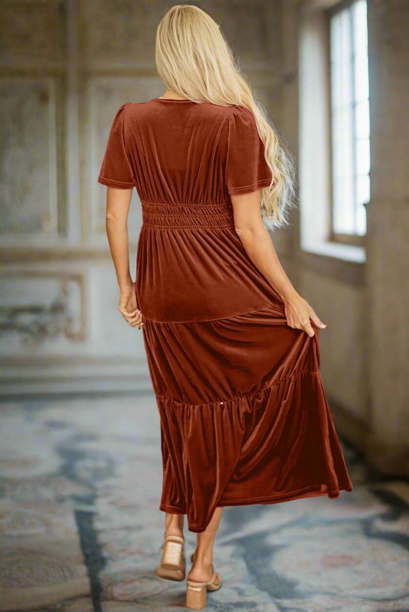 Woman wearing copper velvet maxi dress with tiered design and shirred high waist, viewed from behind in an elegant setting.