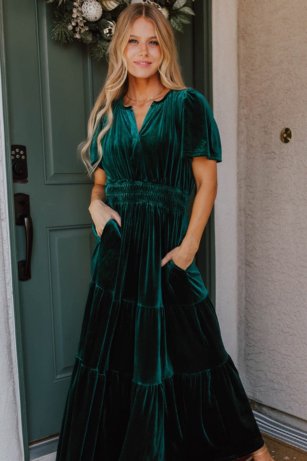 Woman wearing Elegant Emerald velvet maxi dress with short sleeves and tiered design, standing by a green door.