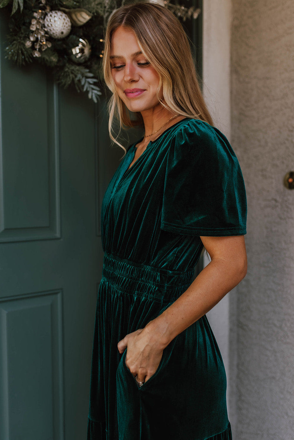 Woman wearing Elegant Emerald velvet dress with V-neck and short sleeves, standing by a green door, showcasing boho style.