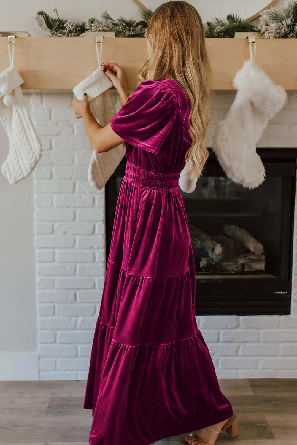 Woman wearing a DARLING DAHLIA Maxi velvet dress with short sleeves and tiered design, standing by a fireplace with stockings.