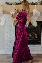 Woman wearing a DARLING DAHLIA Maxi velvet dress with short sleeves and tiered design, standing by a fireplace with stockings.