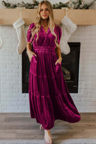 Woman wearing a purple velvet maxi dress with short sleeves and a tiered design, standing in front of a decorated fireplace.