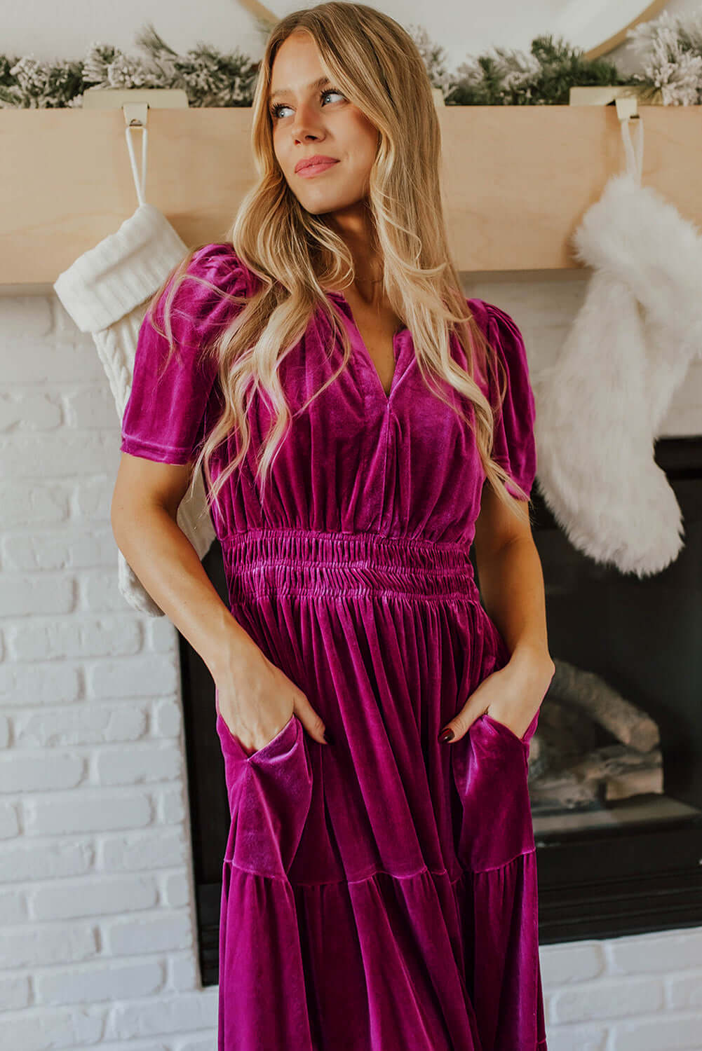 Woman wearing a velvet dress with V-neck and short sleeves standing by a fireplace, showcasing stylish and boho-inspired design.