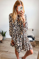 Woman wearing leopard print dress with button front and tiered ruffles, standing in stylish room with plants and cushions.