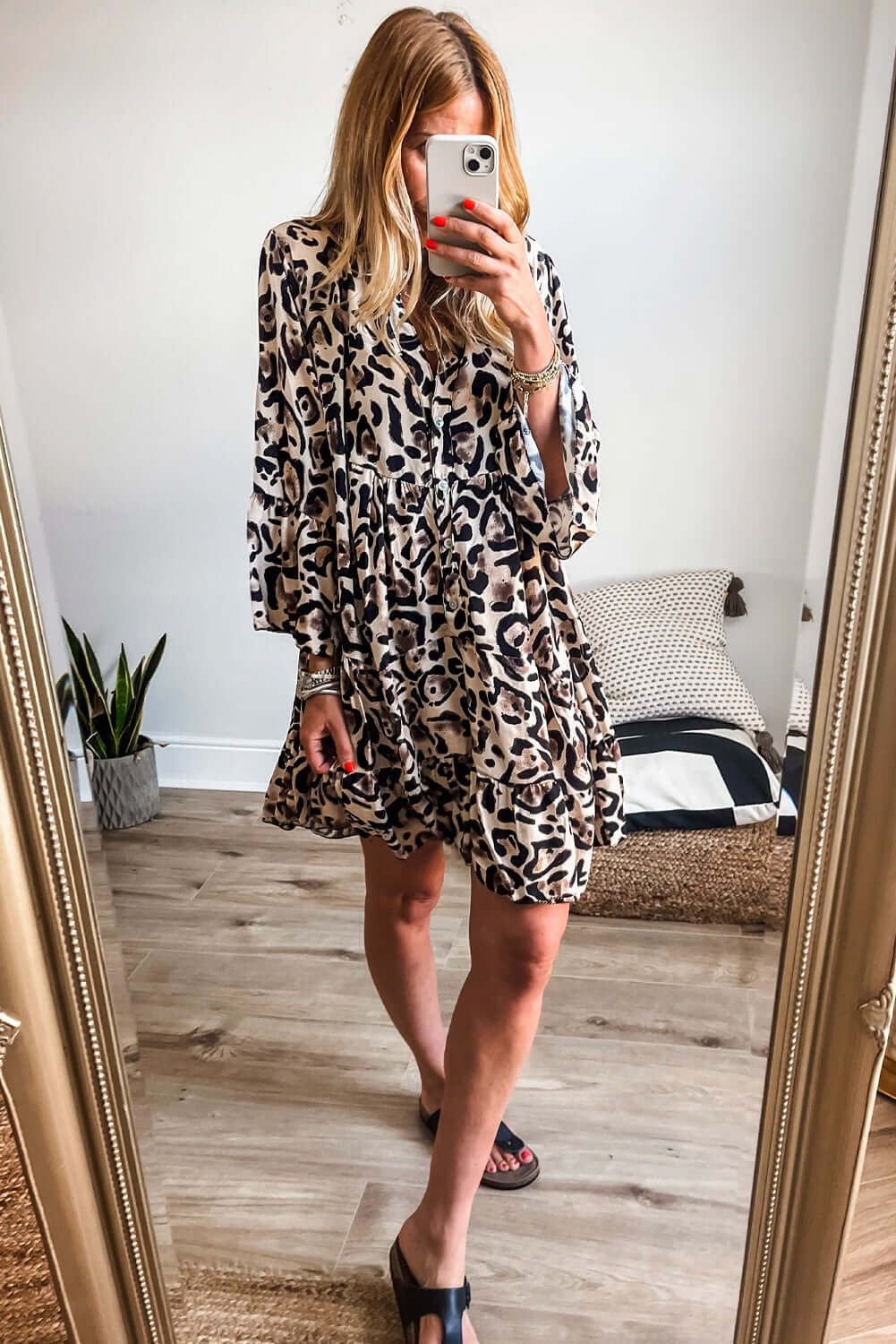 Woman wearing leopard print dress with ruffles, taking a mirror selfie in a cozy room with plants and a stylish decor.