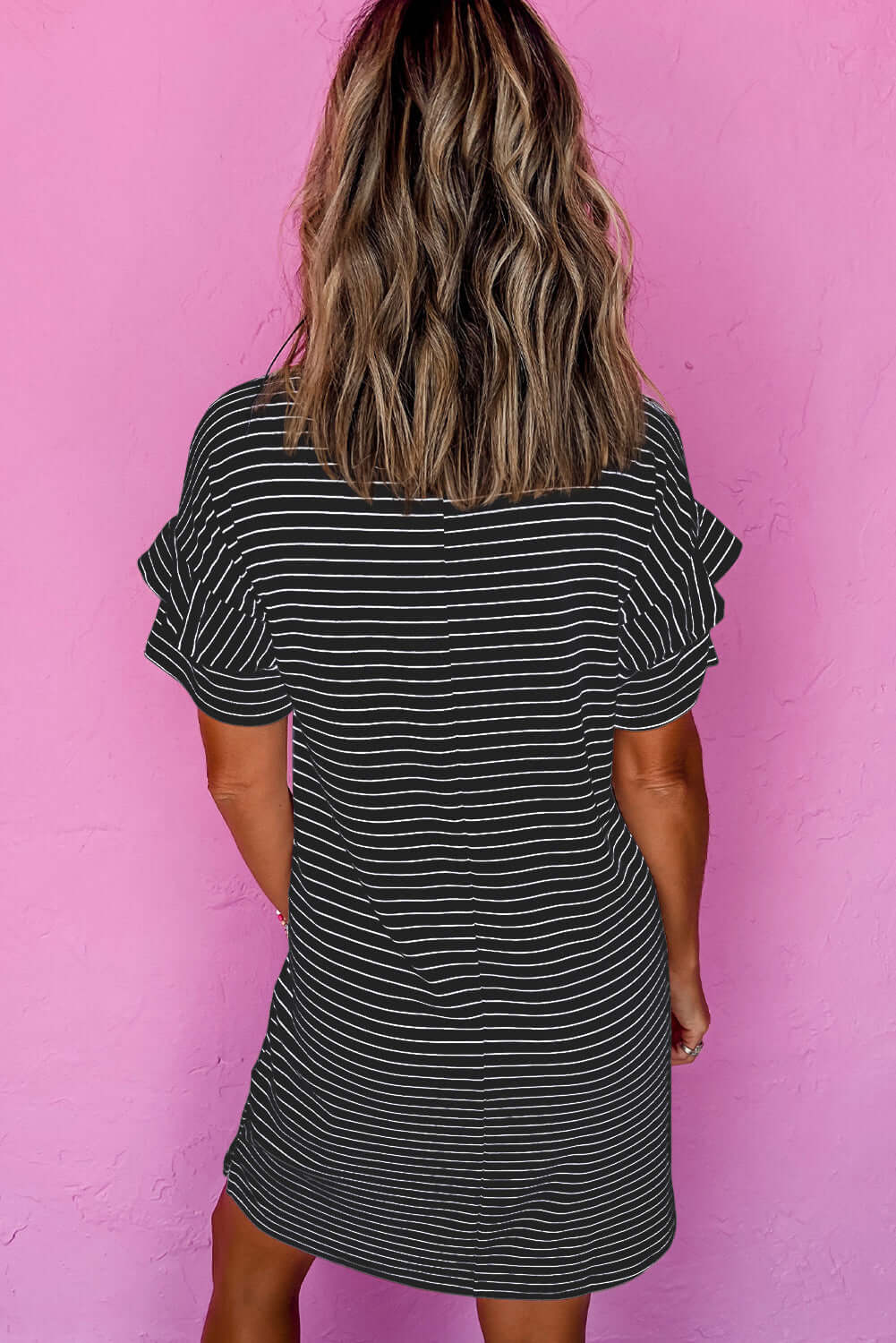 Back view of a woman wearing a black and white striped mini t-shirt dress with ruffle sleeves against a pink background.
