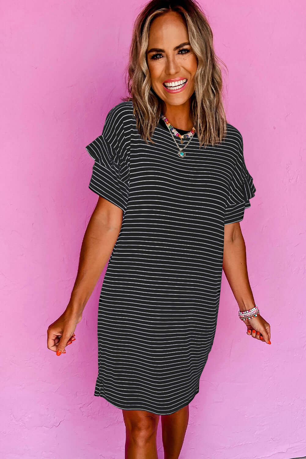 Woman smiling in a black striped mini t-shirt dress with ruffle sleeves against a pink background.