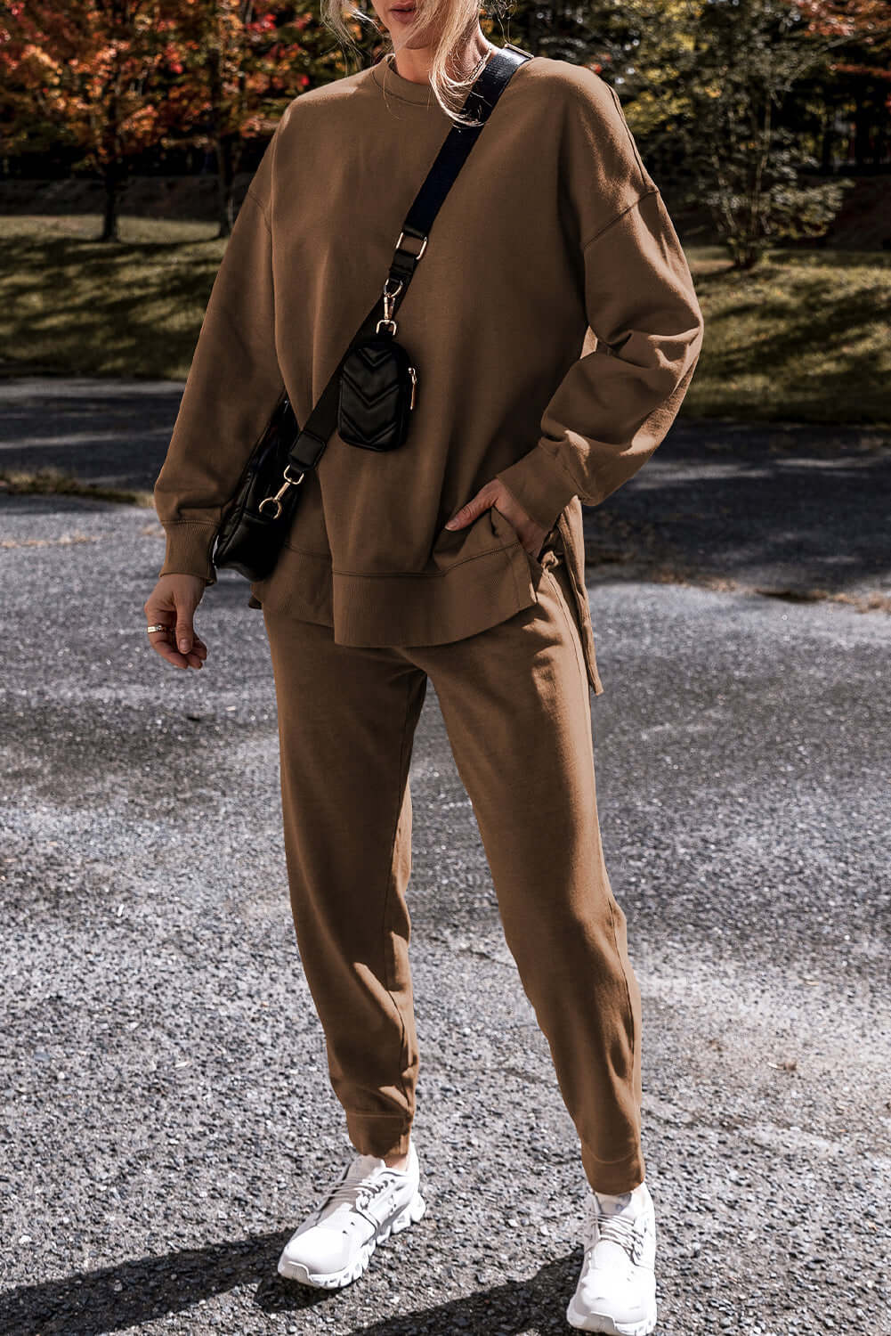 Woman in cocoa high low sweatshirt and lounge pants set, styled with a crossbody bag and sneakers outdoors.