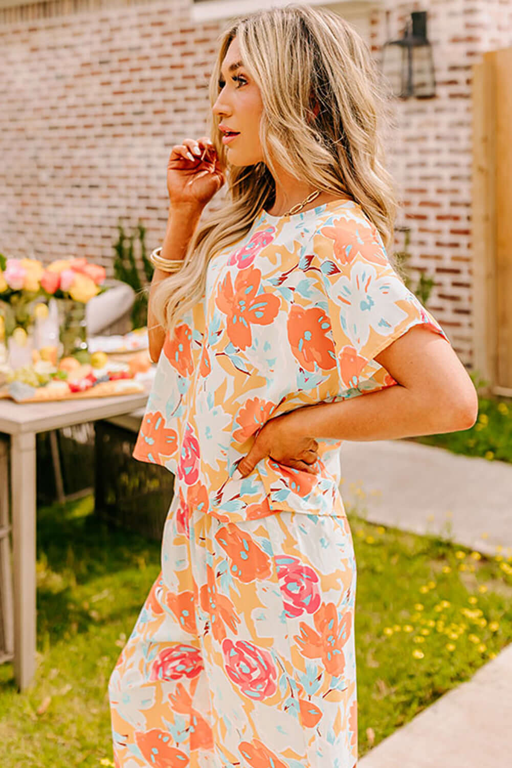 Model wearing SUNSET STROLL PANTS SET, featuring tangerine floral print top and matching wide-leg pants in a garden setting.