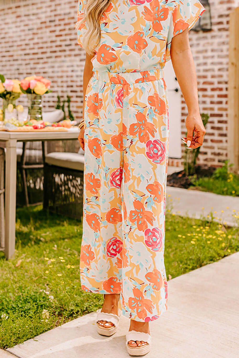 Model wearing tangerine floral printed top and wide-leg pants, stylish outdoor setting with flowers.
