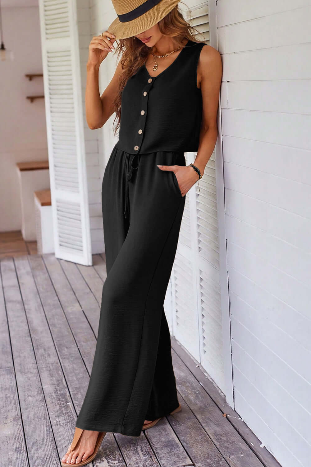Model wearing a chic black buttoned vest and wide-leg pants set, styled with a hat, on a wooden deck.
