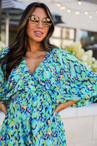 Woman in green and blue abstract print Wave Dancer Romper, wearing sunglasses, smiling outdoors.