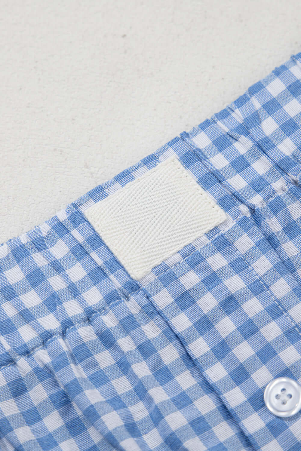 Close-up of blue gingham fabric shorts waistband showing elastic and button details.