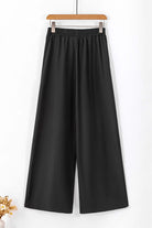 Black wide-leg pants hanging on a wooden hanger, featuring an elastic waistband and flowy design.