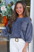 Woman wearing Denim Darlin Top with 3/4 sleeves and chest pocket, standing in front of floral decor.