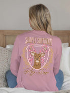 Woman wearing mauve Simply Southern Hey Deer long sleeve t-shirt with deer and bow design, sitting on bed with cozy decor.