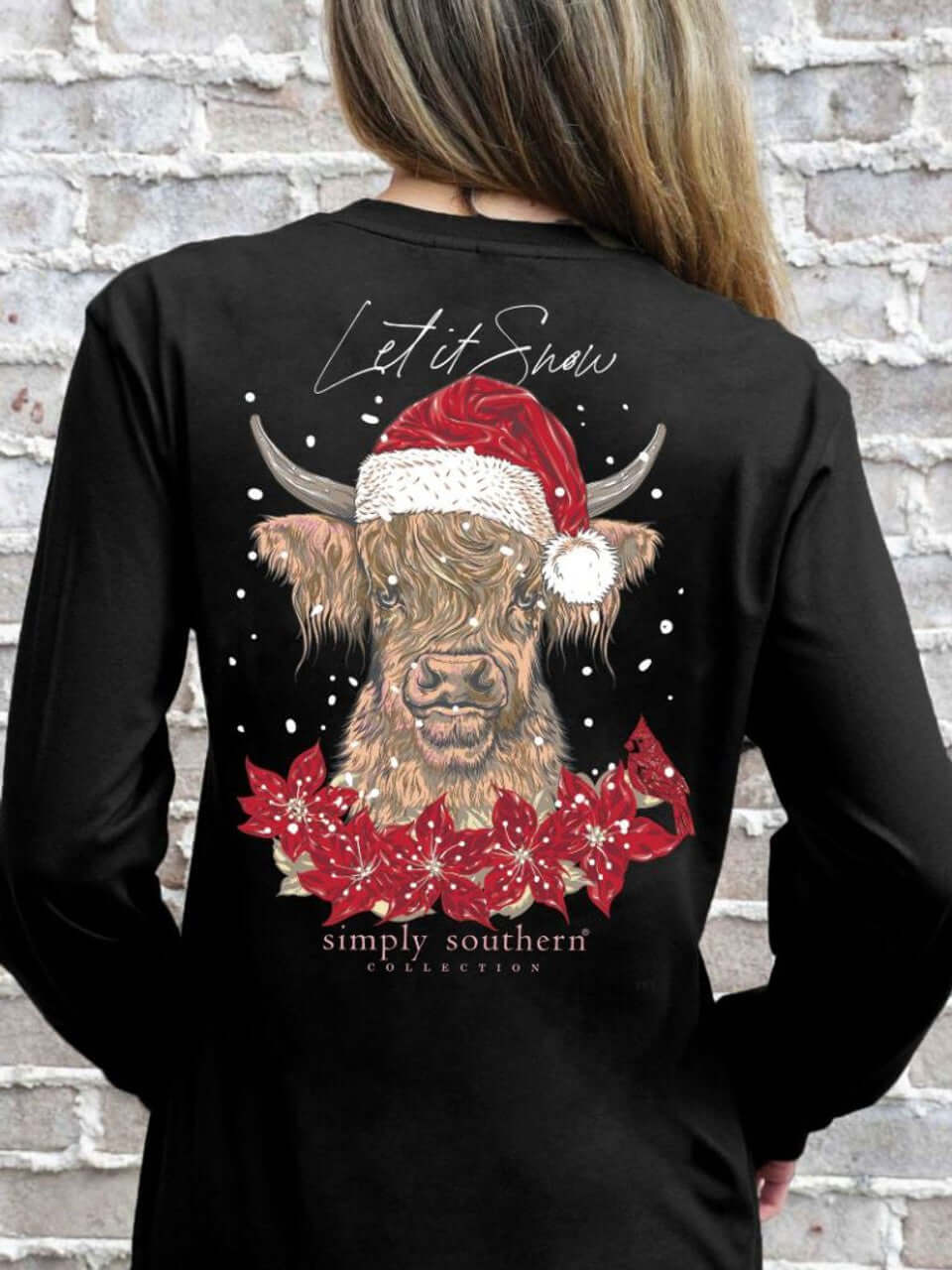 Woman wearing black Simply Southern shirt with a festive highland cow design, featuring a Santa hat and poinsettia wreath, reads "Let it Snow".