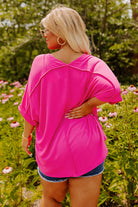 Back view of a plus size woman in a hot pink V-neck top, surrounded by flowers, showcasing relaxed high-low hemline.