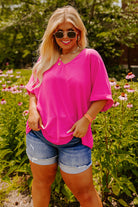 Plus size woman in vibrant hot pink V-neck top and denim shorts, posing among colorful flowers in a sunny garden.