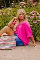 Bright hot pink plus size V-neck top styled with a colorful bag in a floral setting.
