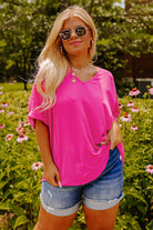 Plus size woman in hot pink V-neck top and denim shorts, surrounded by flowers, showcasing vibrant summer style.
