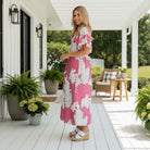 Model wearing JUST BREATHE MAXI DRESS (pink) - Gigio on a porch, showcasing its floral print and flowing design.