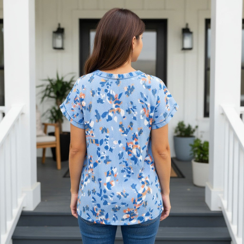 Back view of a woman wearing HELLO YOU TOP (blue) with floral design, standing on a porch.