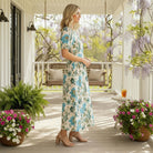 Woman wearing SERENE CHARM DRESS - Gigio with floral print standing on a porch surrounded by flowers.
