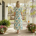 Woman wearing SERENE CHARM DRESS - Gigio with floral print, standing on a porch surrounded by plants and flowers.