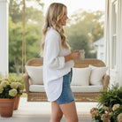A woman in a white shirt enjoys a drink while wearing CAMRYN SHORTS - Judy Blue on a porch with plants.