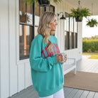 Woman smiling in LULU MAC Aqua with Coral Puff Logo Crew sweatshirt, holding a cup on a porch.