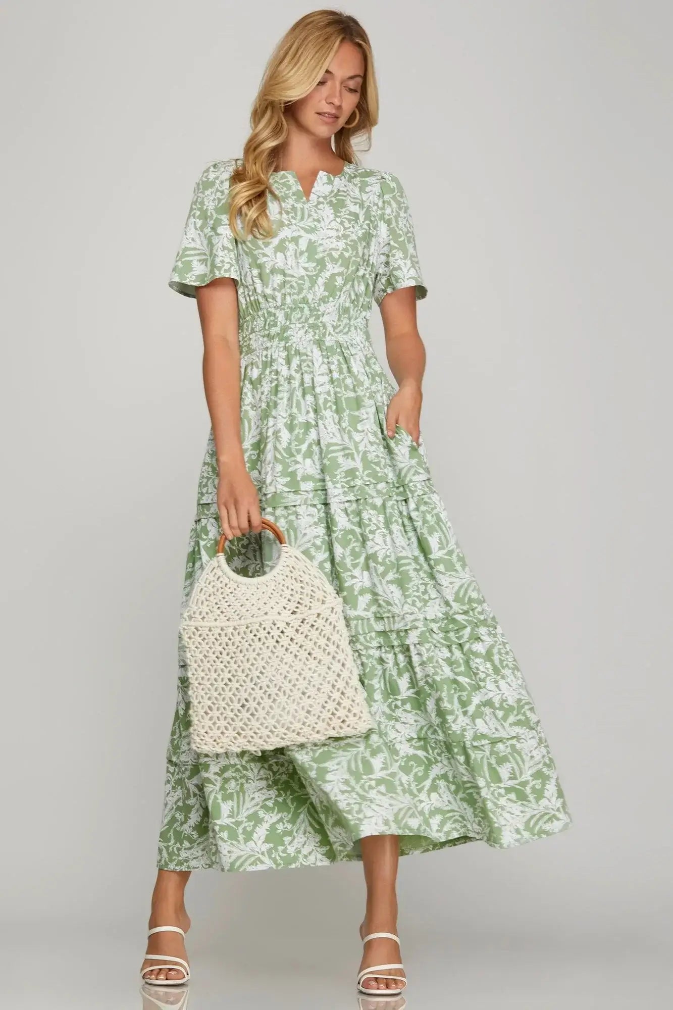 Woman in sage floral maxi dress with flutter sleeves and side pockets, holding a woven bag.