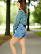 Model showcasing JUDY BLUE Cassidy Shorts in a park setting, styled with a floral top, highlighting casual summer fashion.