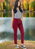 Woman wearing Judy Blue Bridget Joggers in crimson with tank top, standing by a lake with autumn trees in the background.