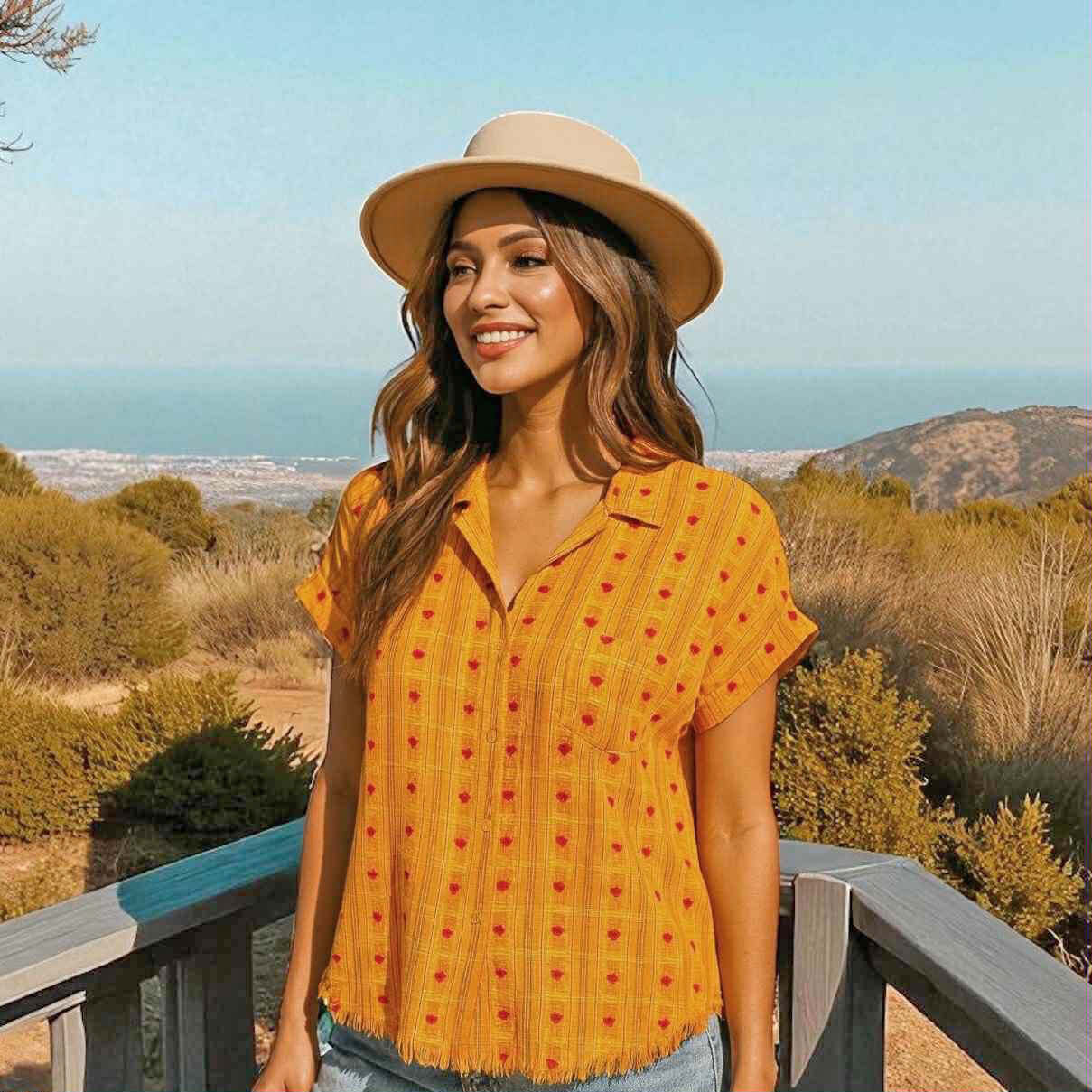 Bright yellow button-down blouse with heart motifs, styled with a hat against a scenic outdoor backdrop.