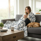 Cozy woman wrapped in a SNOWY MOUNTAIN PLUSH THROW BLANKET with a dog on a sofa, enjoying a warm drink.