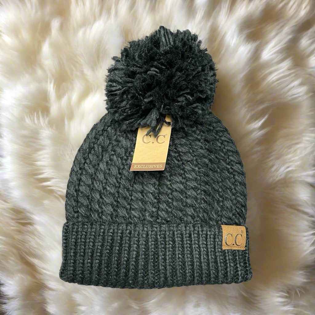 Black cable knit beanie with pom pom and suede CC patch on faux fur background.