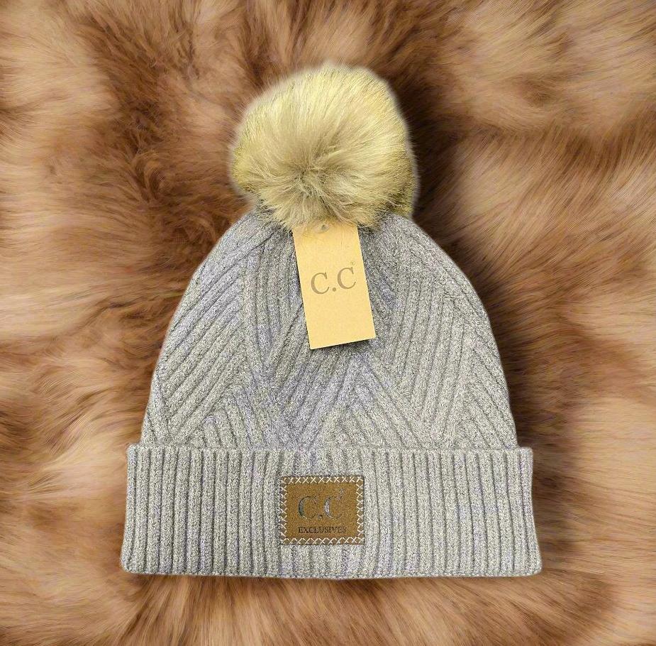 Light gray pom pom beanie with faux fur and diagonal pattern, featuring a C.C. tag, placed on a fur-like surface.