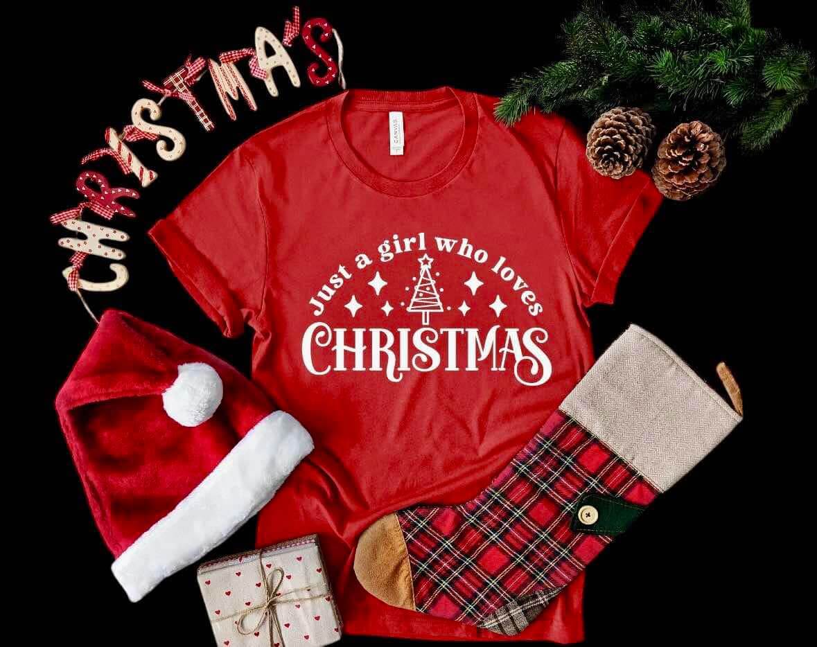 Cozy red Christmas tee with festive graphics, surrounded by holiday decor including a Santa hat, stocking, and pinecones.