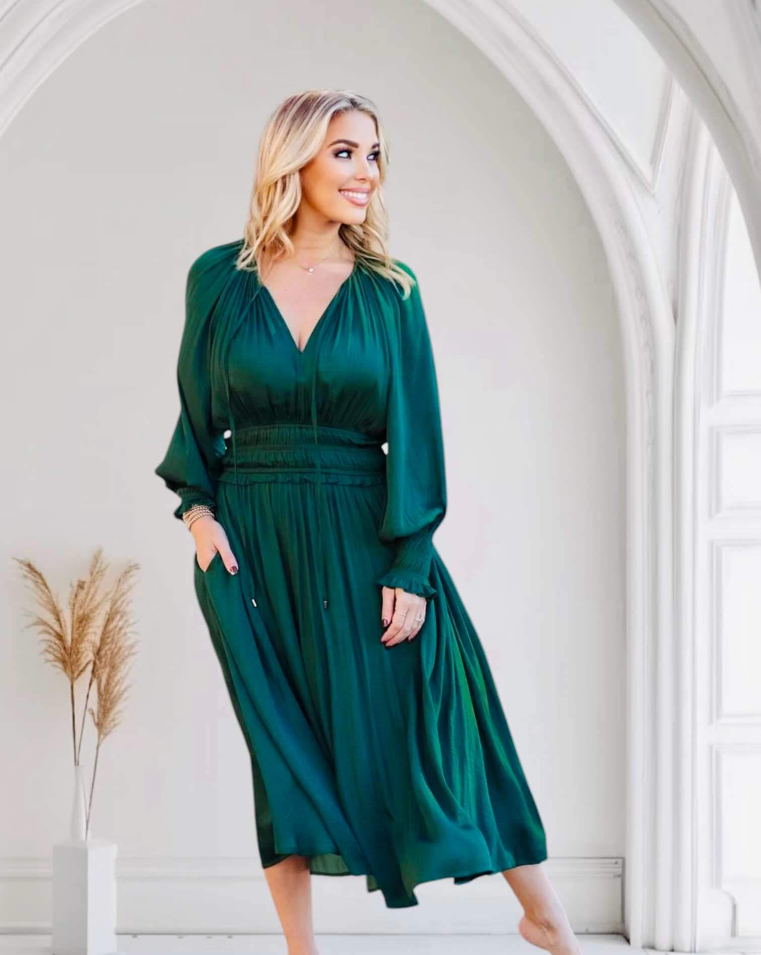 Woman modeling dark green midi dress with smocked sleeves and waist by Pinch, standing elegantly in an archway with pampas grass decor.