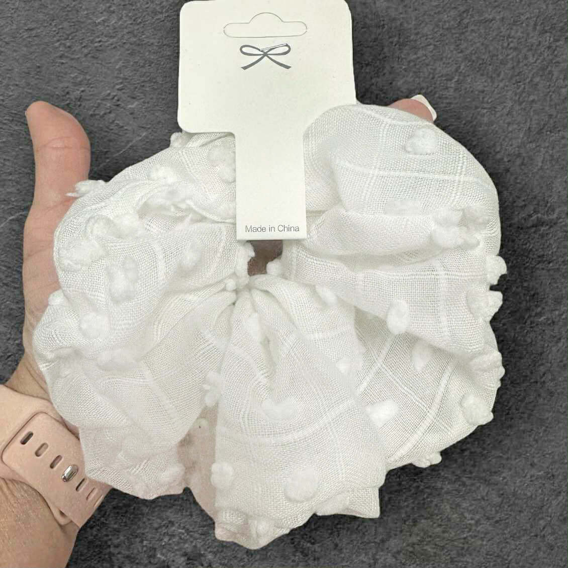 White pompom textured scrunchie held in hand; stylish hair accessory, perfect for tying back hair.