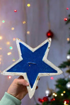 Hand holding a blue star-shaped dough bowl candle with rustic charm, featuring a mahogany teakwood scent, against a festive background.