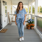 Smiling woman wearing the HELLO YOU TOP (blue) with floral design, paired with jeans and casual shoes, on a porch.