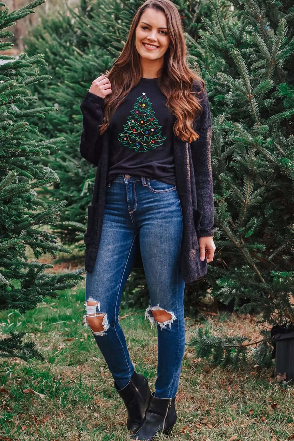 Woman in a studded Christmas tree t-shirt, styled with ripped jeans and a cozy cardigan, surrounded by pine trees.