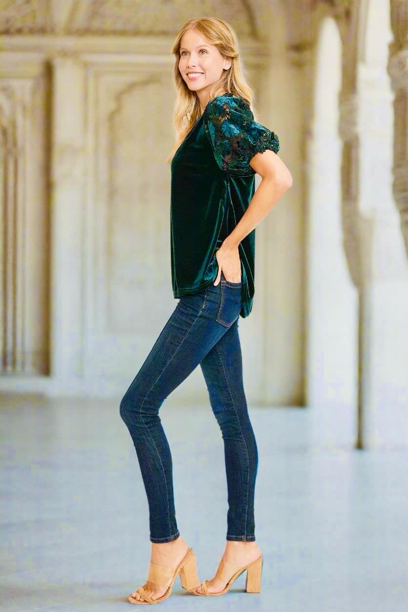 Woman wearing Umgee QUIET JOY Velvet Top in dark teal, featuring lace sleeves, paired with jeans and heels, standing in elegant setting.