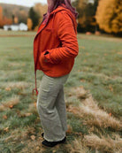 Woman wearing an Autumn Sunrise Jacket in ginger shade, standing in a grassy field with autumn trees in the background.